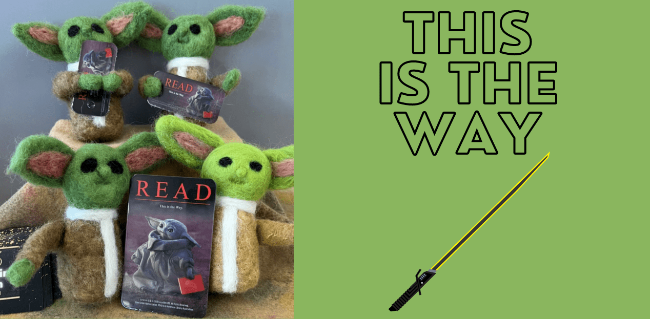 Felted baby yoda dolls holding baby yoda library cards.  Also, a lightsaber with the text This is the Way.